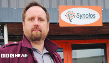 Barry Ingleton, a man with brown hair and a full beard, looks at the camera. He is wearing a maroon coat with an upturned collar over a grey shirt. Behind him is a sign on the front of a building reads "Synolos" next to a stylized symbol representing a tree.