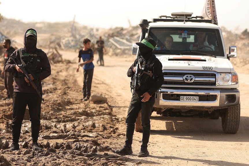 Two men wearing face coverings and green headbands, holding rifles, escort a jeep through rubble.