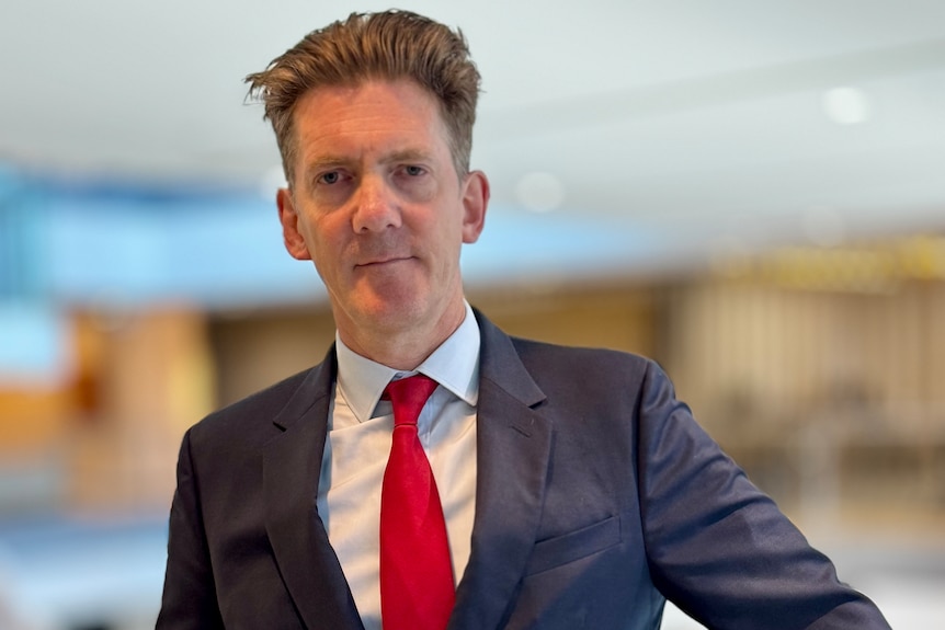 A tall middle-aged man in a dark suit and red tie looks at the camera