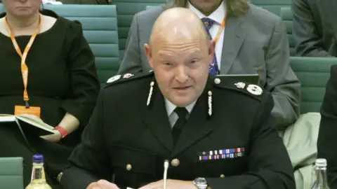 UK Parliament Former Chief Constable Craig Guildford sits at a desk while wearing a black police uniform with a medal ribbon on it. Several people are sitting on green chairs behind him.