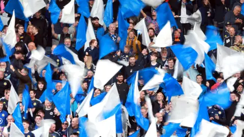 PA Media Football fans wave blue and white Scotland flags while watching the Scotland v Finland international friendly being played in Glasgow in 2024.