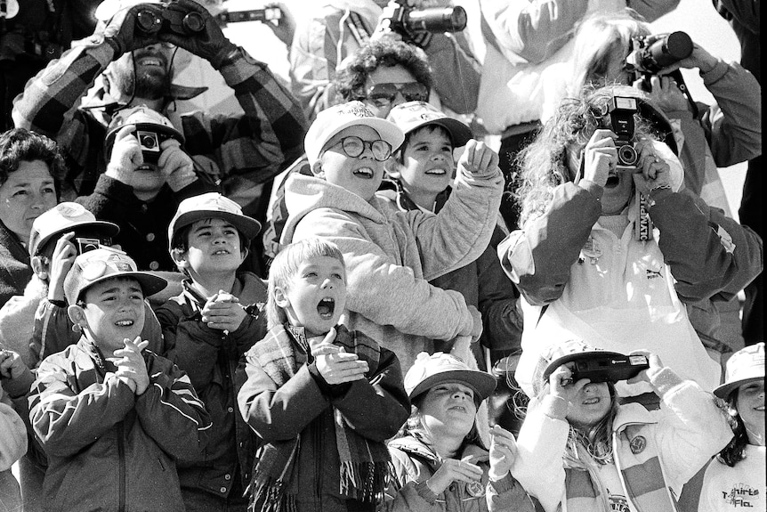 Black-and-white photo showing people in tiered rows, some with cameras, some clapping, smiling excitedly and looking upwards.