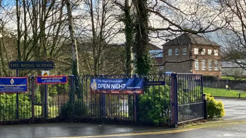 The tree-lined entrance to the Royal School, Armagh. Blue metal railings line a pedestrian and vehicular entrance to the site. There is a car park and a modern red-brick building in the background.