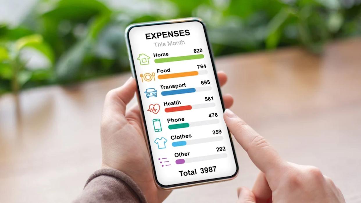 A person holds a smartphone displaying a colorful expense tracker app with categories for home, food, transport, health, phone, clothes, and other, showing totals for each and a monthly total of 3987.