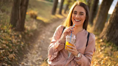 Getty Images A young woman walks along a rural path lined by trees. She's holding an iced drink in a clear takeaway cup and smiling.