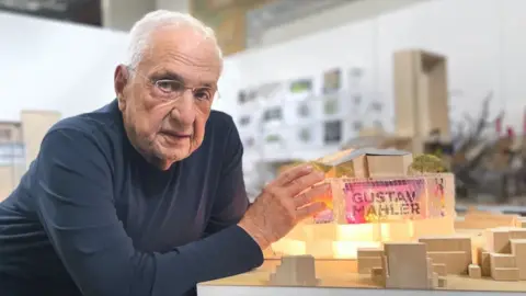 Wimbledon Concert Hall Trust A person wearing a dark long-sleeve shirt is leaning on a table, presenting a detailed architectural model, featuring illuminated sections, miniature trees, and geometric structures in a modern studio setting.
