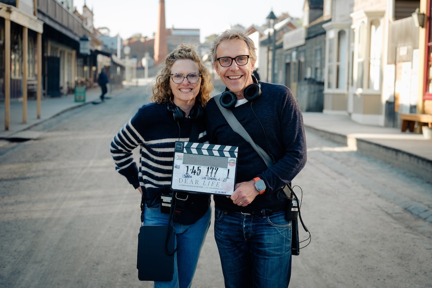 Robyn Butler and Wayne Hope, both middle-aged, smile on a street in Ballarat, holding a clapperboard that reads "Dear Life".
