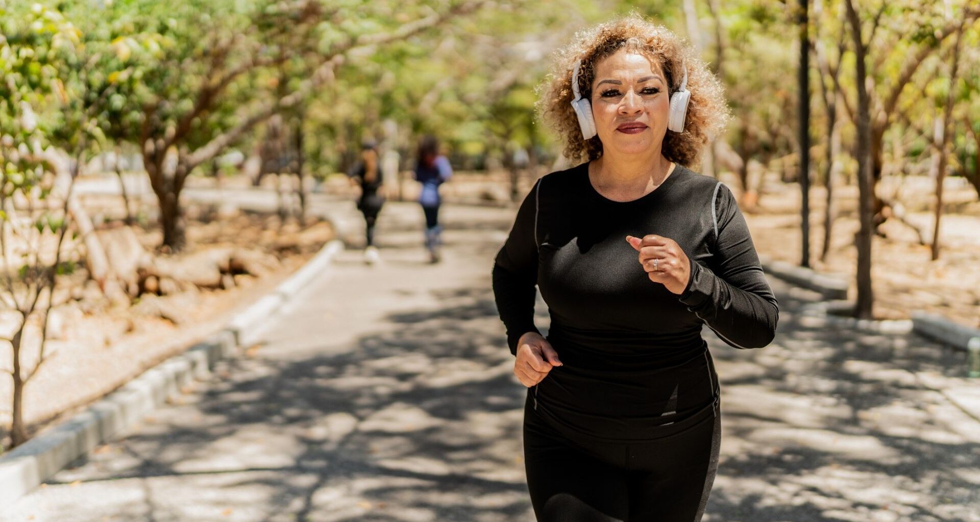 woman dressed in black sleeved top and leggings running outside with white headphones. she's on a path with trees and other people blurred in the background