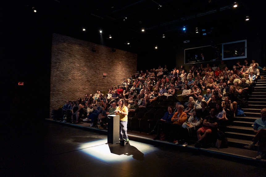 A person stands at a plinth with a controller, the camera is looking towards an auditorum of people in seats