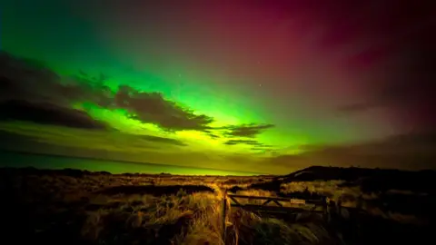 Alan Kneen Landscape shot of the Northern Lights taken from a field. The sky is dark red bursting into bright green and yellow.