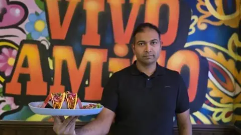 Rajendra Vikram Kupperi Rajendra Vikram Kupperi wearing a black short-sleeved shirt is holding a large white plate with three vibrant tacos. Each taco is filled with colorful ingredients, including shredded lettuce, diced tomatoes, and possibly meat, topped with bright pink pickled onions. The background features a bold, colorful mural with swirling patterns and large orange letters spelling “VIVO AMIGO” against a mix of blue, yellow, and pink designs.