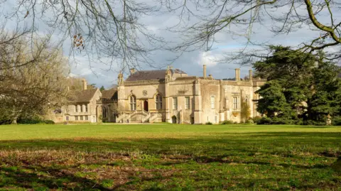 Getty Images Lacock Abbey in Wiltshire. The building is old and beige. There are trees and green fields around it.