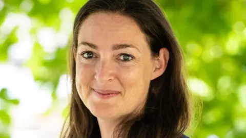 RNID Crystal stands outdoors against a backdrop of bright green foliage. She is wearing a dark blue short-sleeved top with a simple, modest design. Her hair is long, straight, and dark brown, falling over the shoulders.