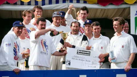Getty Images Essex cricketers wearing their whites and celebrating winning the County Championship in 1992. They are holding champagne and a large cheque for £46,000.