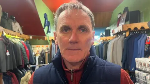 BBC A man in a blue gilet and an orange fleece stands in the middle of men's clothes shop. A number of suits and items are on rails behind him.