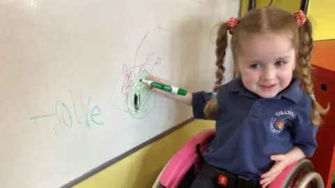 Ann Reel Hollie at school writing on the whiteboard. She has her red/brown hair in pigtails. 