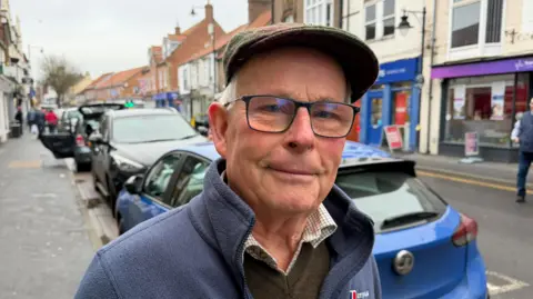 Becki Bowden / BBC A man wearing a flat cap and black-rimmed glasses stands on pavement beside a high street in a market town. He is wearing a blue fleece, brown jumper and white checked shirt. Cars are parking beside him at the side of the road, which is lined by shops.