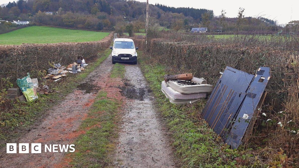 A country lane is pictured with mattresses, building waste and metal sheets piled on the grass verges on either side. A white van is parked on the road in shot.