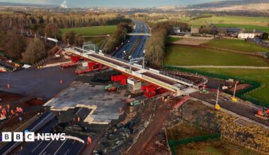 An aerial view of the new bridge being positioned over the M6. It is a light grey with bright red supports underneath rolling it into place.