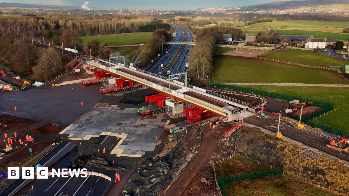 An aerial view of the new bridge being positioned over the M6. It is a light grey with bright red supports underneath rolling it into place.