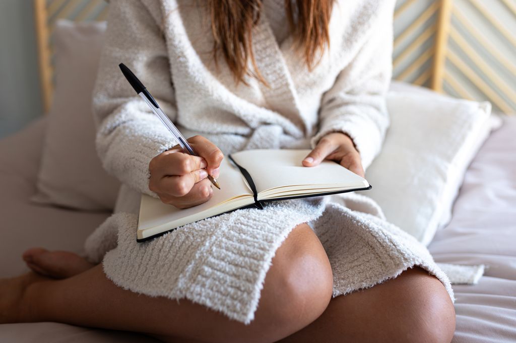 Young woman writing in notebook while sitting on bed in bathrobe