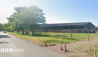 A Streetview image of a road with a large industrial building to the right. A track off the road is blocked by wire link fence and a few orange cones.