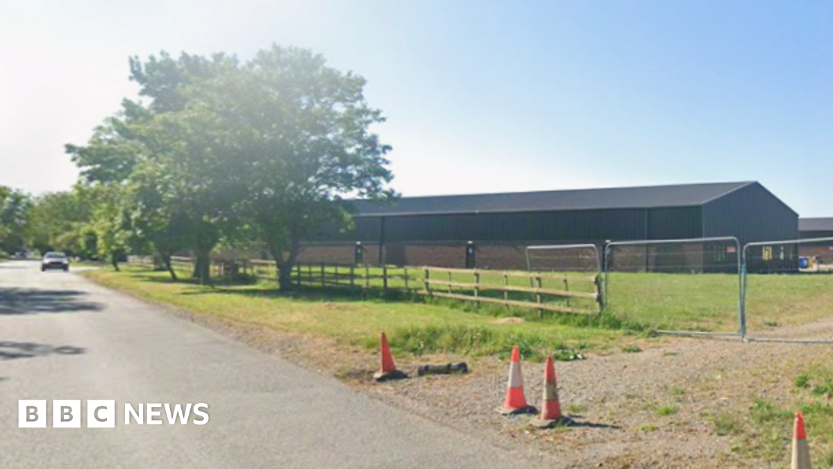 A Streetview image of a road with a large industrial building to the right. A track off the road is blocked by wire link fence and a few orange cones.