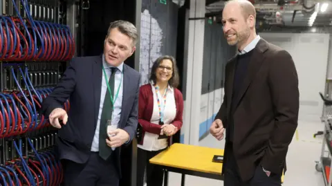 PA Media Prince William looks on as a man in a suit points out elements of the Isambard super computer. There are racks of blue and red cables, all looped around neatly and plugged in. A woman wearing a lanyard looks on cheerfully. 