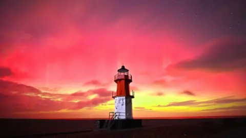 Nigel Fairclough Landscape shot of the northern lights from the Point of Ayre. The lighthouse, painted red and white, sits under a bright red, yellow and blue sky.