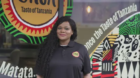 Photograph of Justina John stood outside of her store, Onja. She has long black curled hair, brushed around the side, on her left shoulder. He has brown eyes and smiles at the camera. The restaurant window reads: "Taste of Tanzania" alongside vibrant tribal prints. 