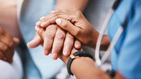 Getty Images A stock image of a nurse holding the hand of a patient. The nurse is wearing a black watch, and her blue uniform is blurred to the right.
