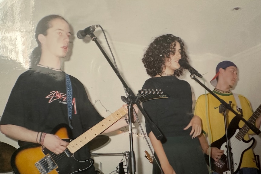 Dressed in head to toe black, Yasmine Ghoniem sings at a microphone, two guitar players either side of her.