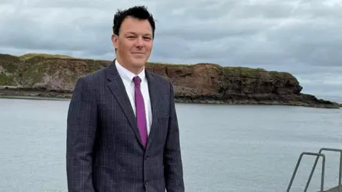 Facebook A dark-haired man with a blue jacket, white shirt and purple tie faces the camera with a seascape in the distance