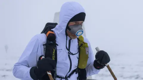 David Murch/Montane Spine Race A man in a white waterproof jacket with the hood up, standing in a snowy landscape. He is also wearing a backpack and a mask over his face, and is carrying walking poles.