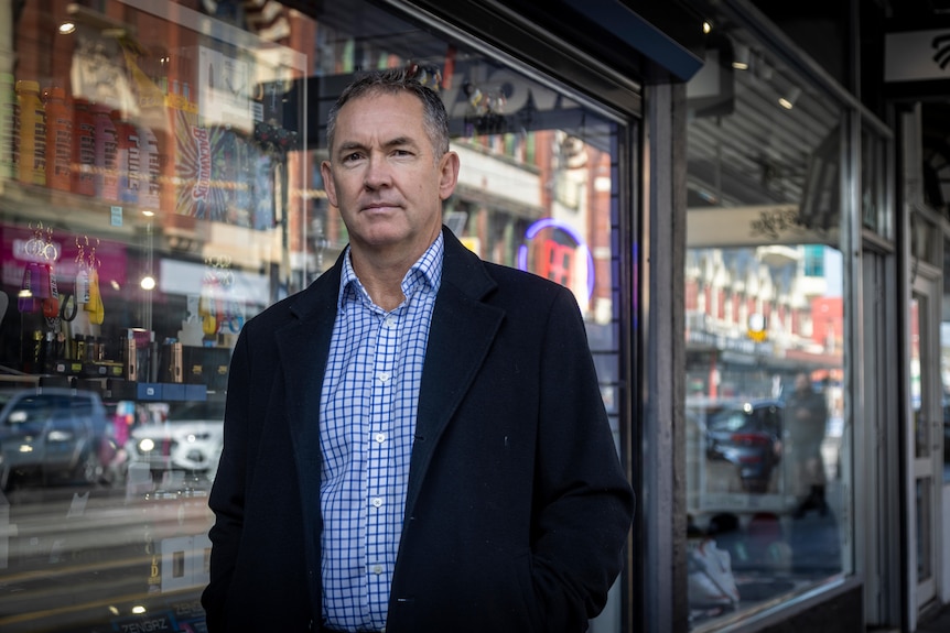 Medium-close-up shot of a man wearing a black jacket and blue checked shirt in front of a shop window.