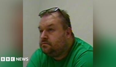 A man with grey hair and stubble is wearing a green t shirt and his glasses are on top of his head. He is sitting in front of a white wall.