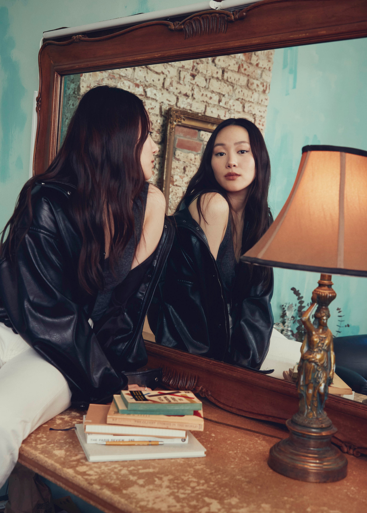 Woman in black leather jacket sits on a vintage wooden desk, looking at herself in a large framed mirror; books and a decorative lamp are on the desk, with exposed brick and turquoise walls in the background.