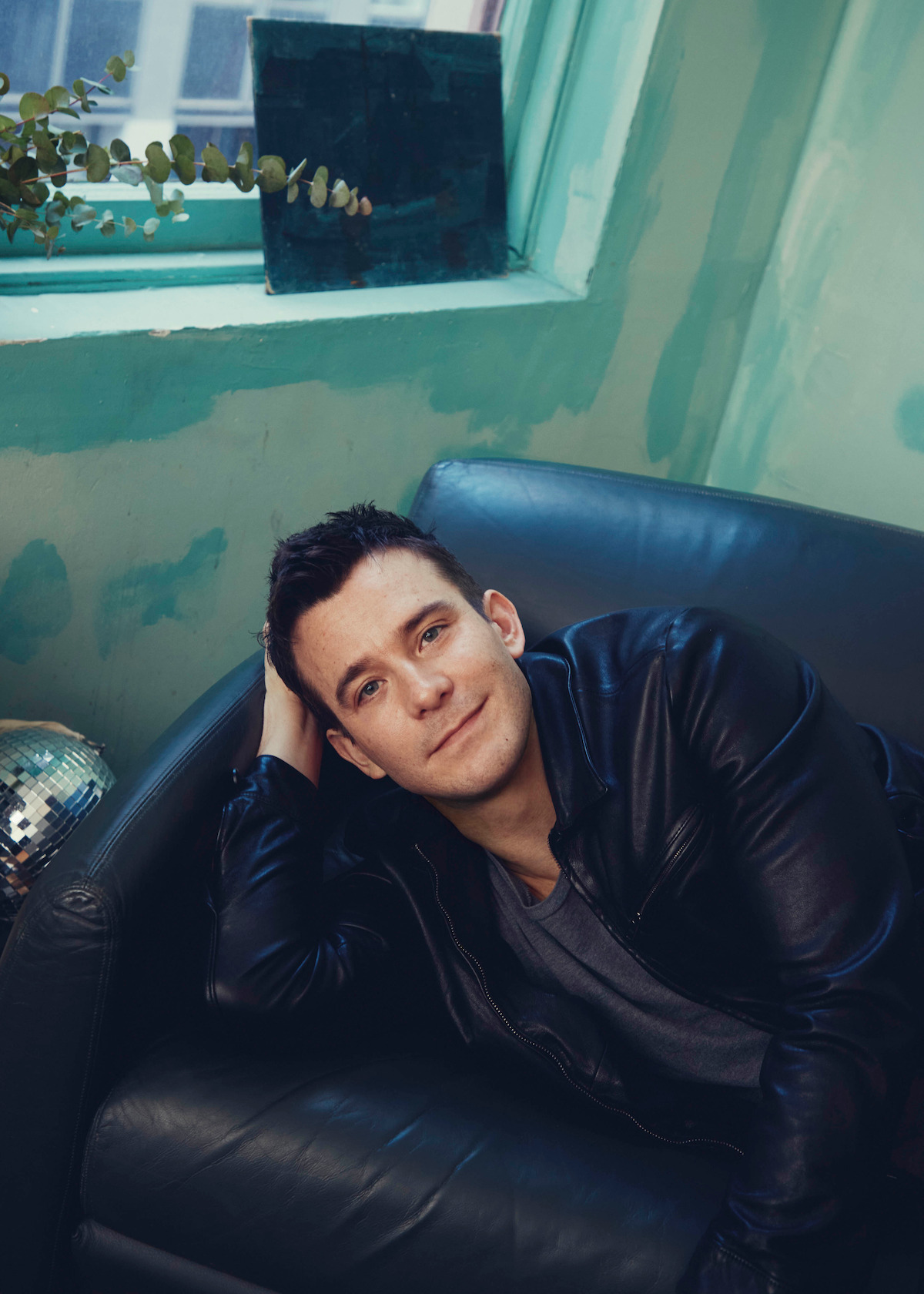 Man in black leather jacket relaxing on a black leather chair in a softly lit room with green walls, next to a window with plants and a disco ball partially visible.
