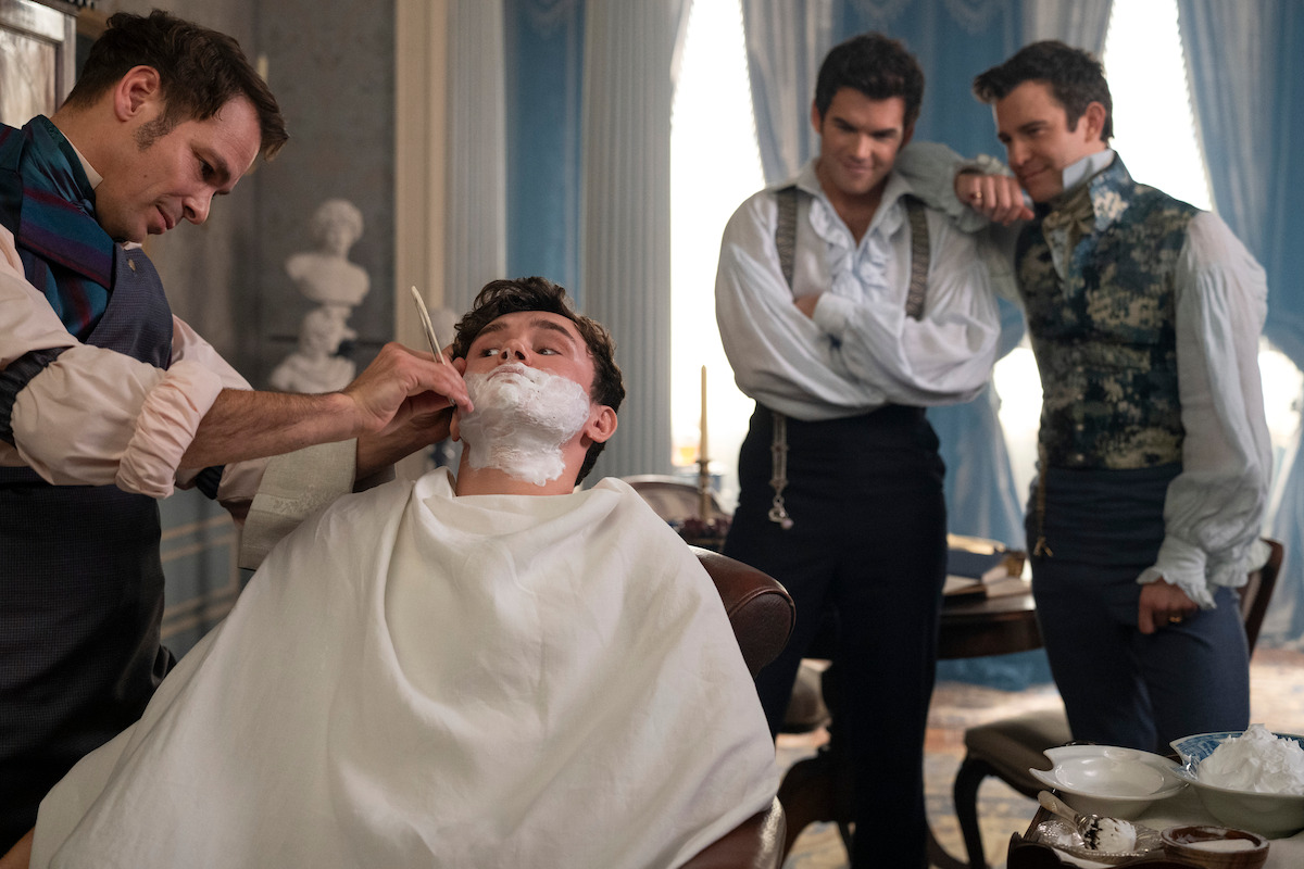 A man gets a traditional shave in an elegant, classical room as two well-dressed men watch and smile.