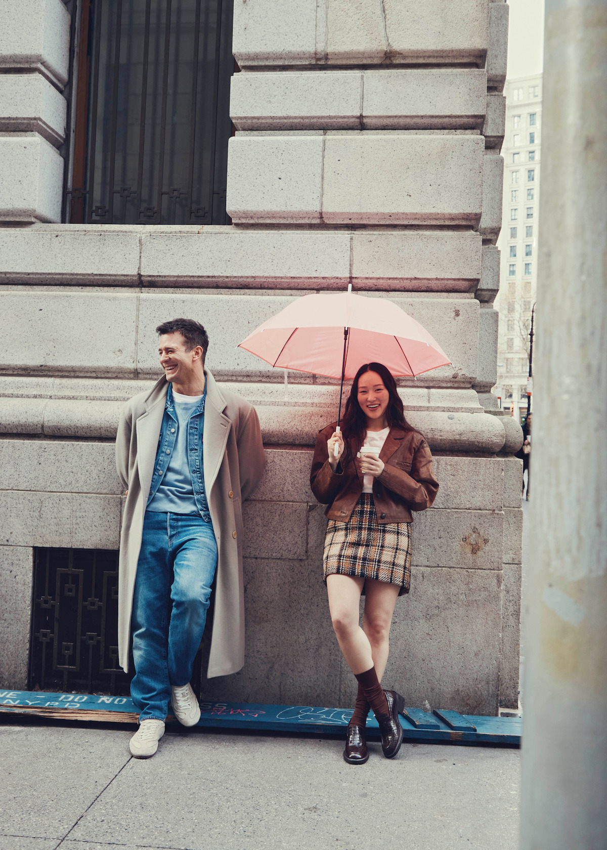 A man in a coat and a woman holding a pink umbrella and a drink stand smiling against a stone building in an urban city setting, suggesting a casual and relaxed atmosphere.