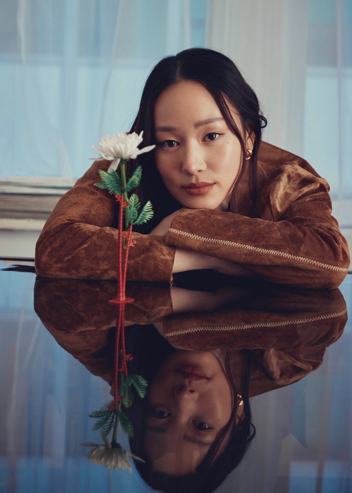 A woman in a brown jacket rests her arms on a piano, gazing at the camera with a white flower nearby; her face and the flower are reflected on the glossy surface in a softly lit, indoor setting with sheer curtains.
