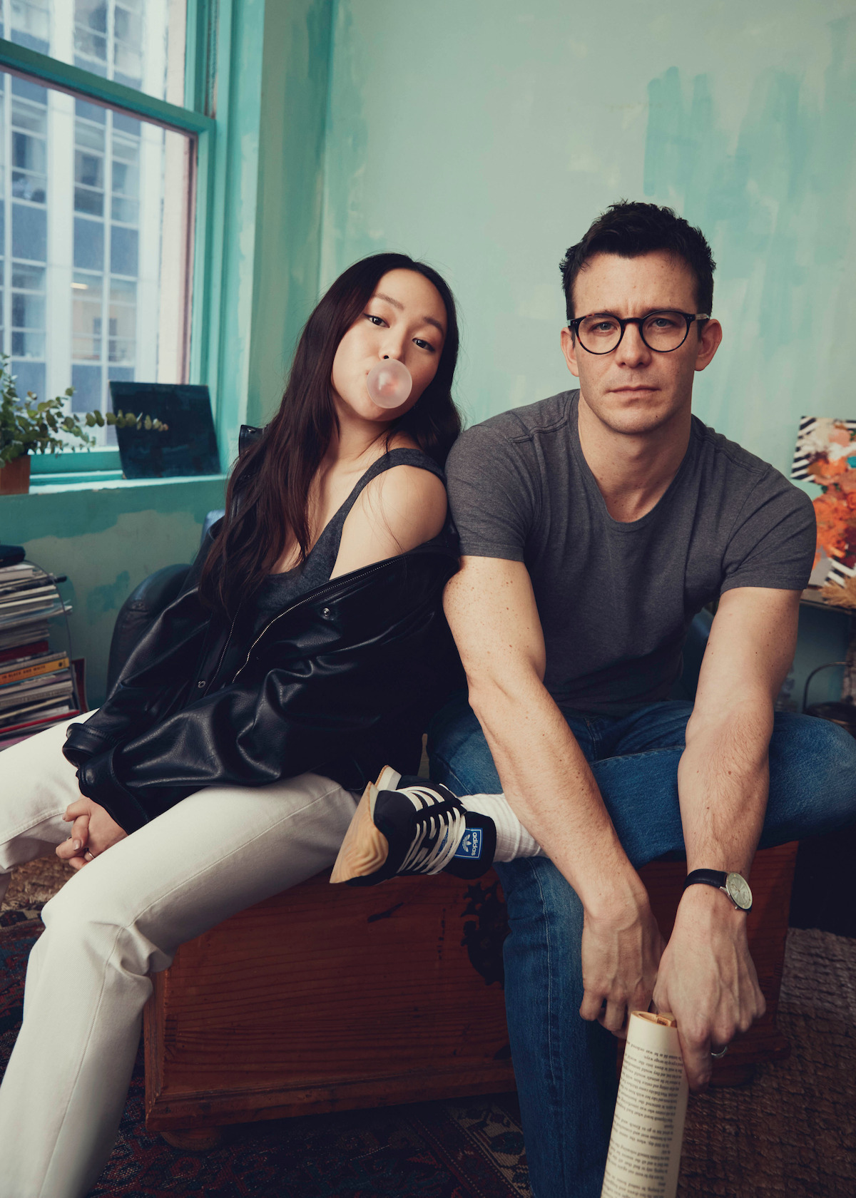 A woman blowing bubble gum and a man with glasses sit together in a casual, artistic room with green walls, books, a plant, a window, and a patterned chair, creating a relaxed, modern atmosphere.