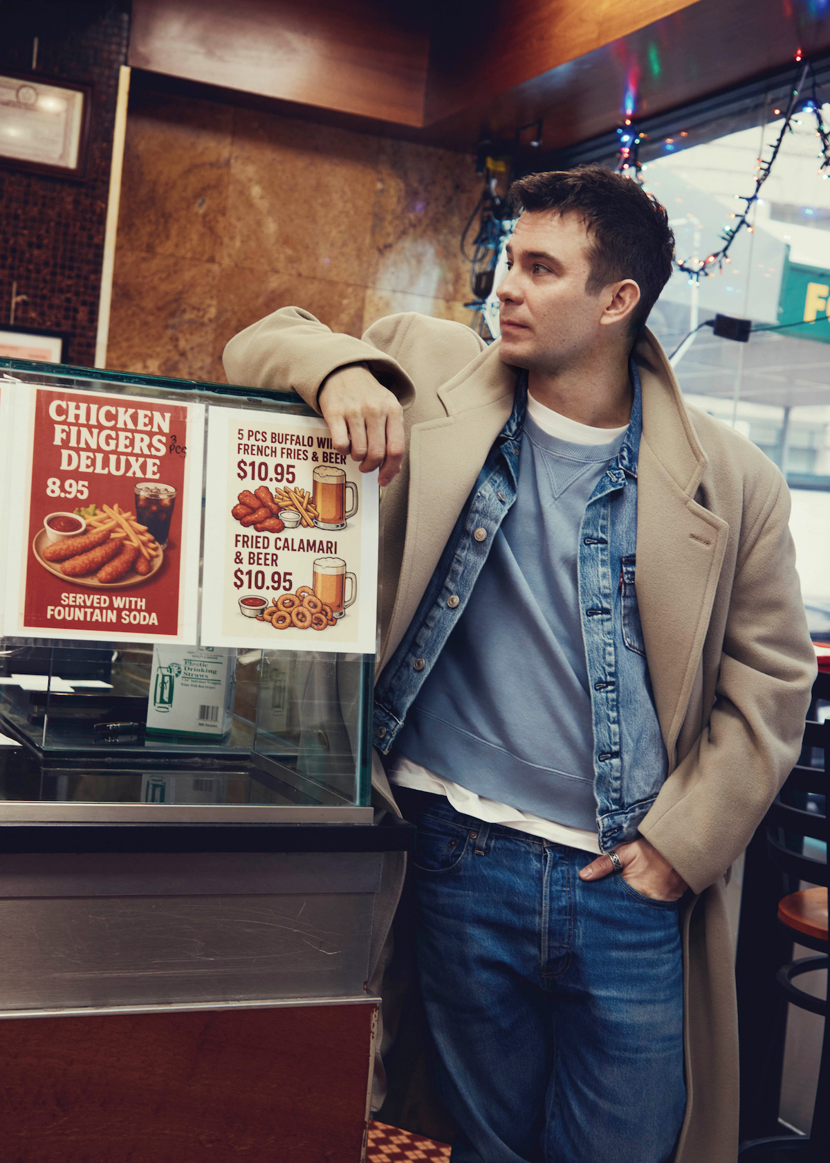 Man in casual blue and white outfit with beige coat stands inside a diner, leaning on a counter near menu signs for chicken fingers and calamari, with a casual, urban interior and string lights visible.