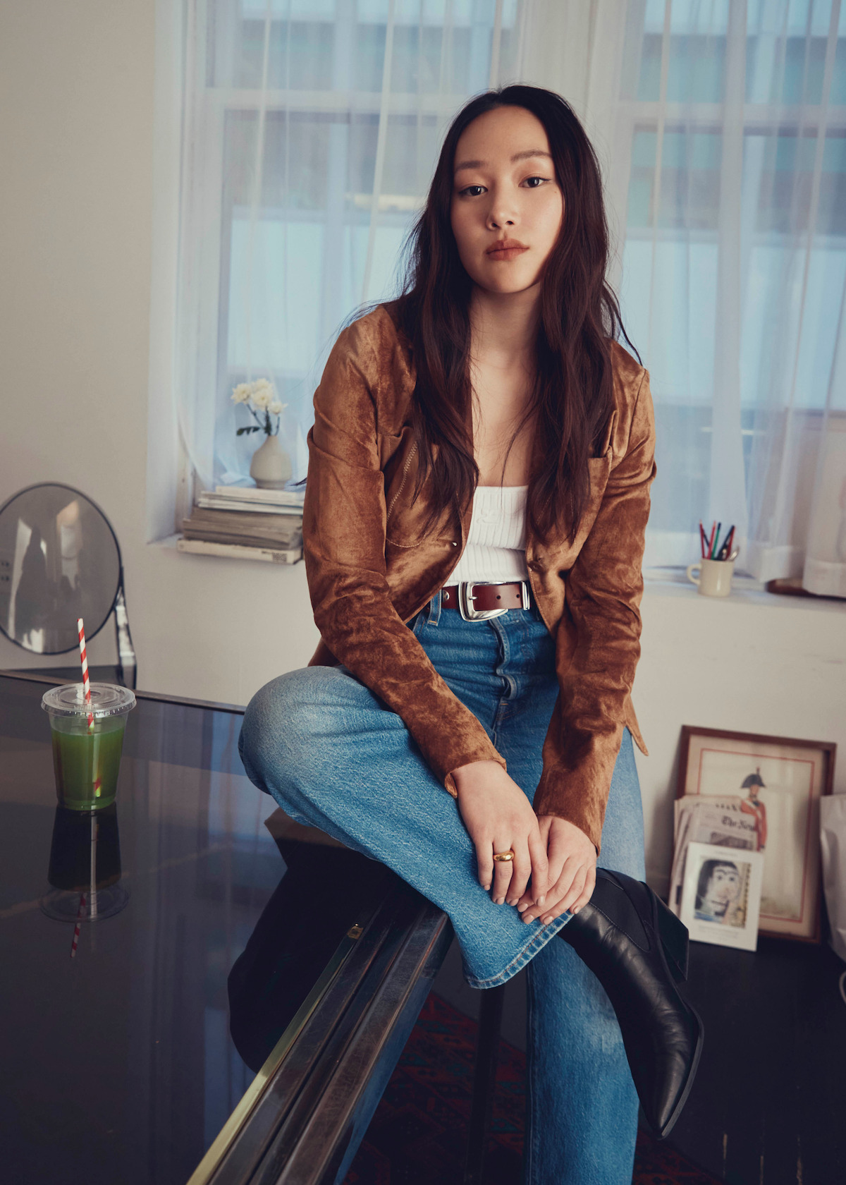 Woman with long brown hair sits on a table in a cozy room with large windows, plants, framed photographs, and a green drink with a striped straw on the desk, creating a relaxed, artistic atmosphere.