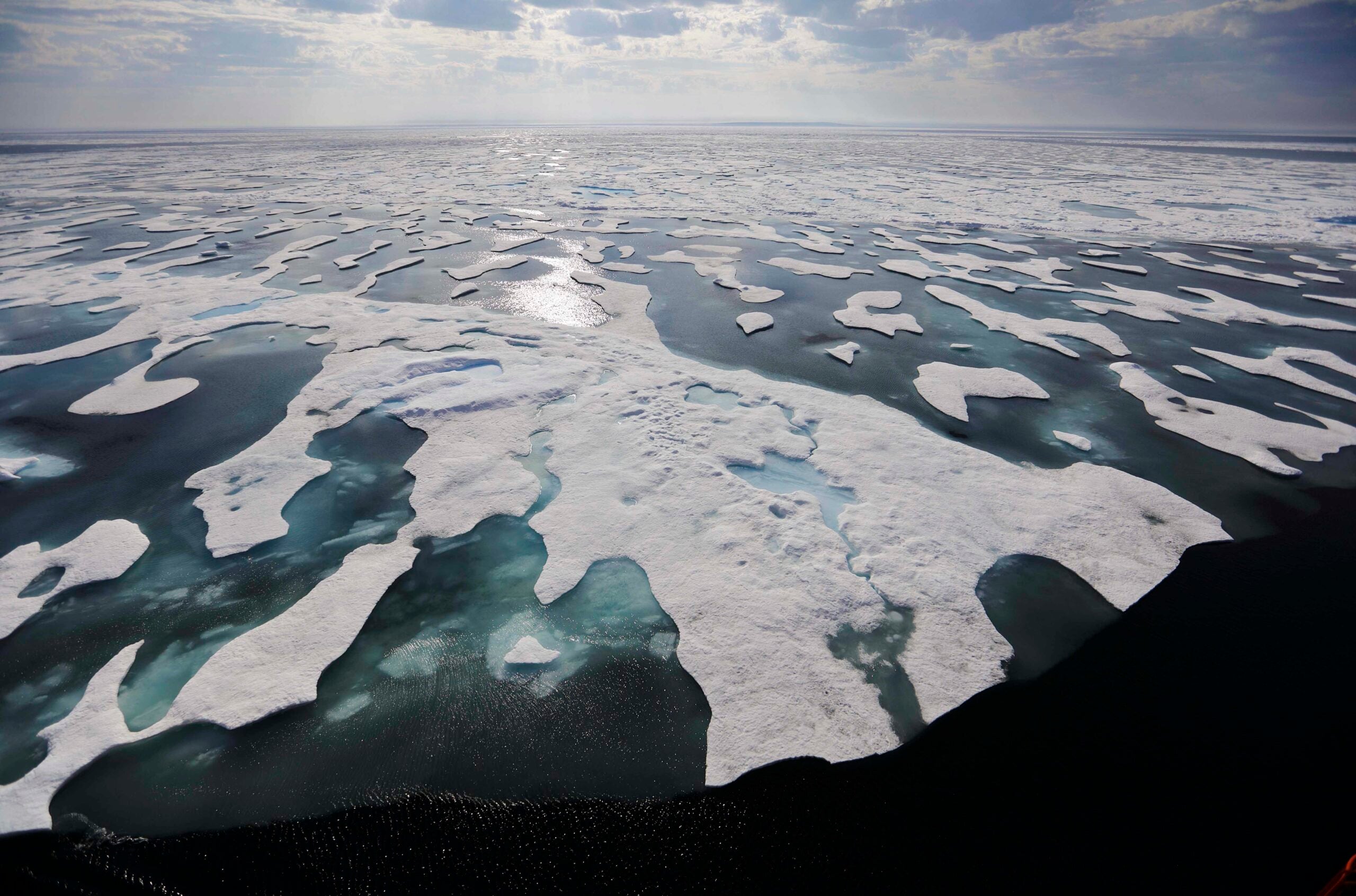 Aerial view of melting sea ice with patches of open water in the Arctic Ocean under a partly cloudy sky.