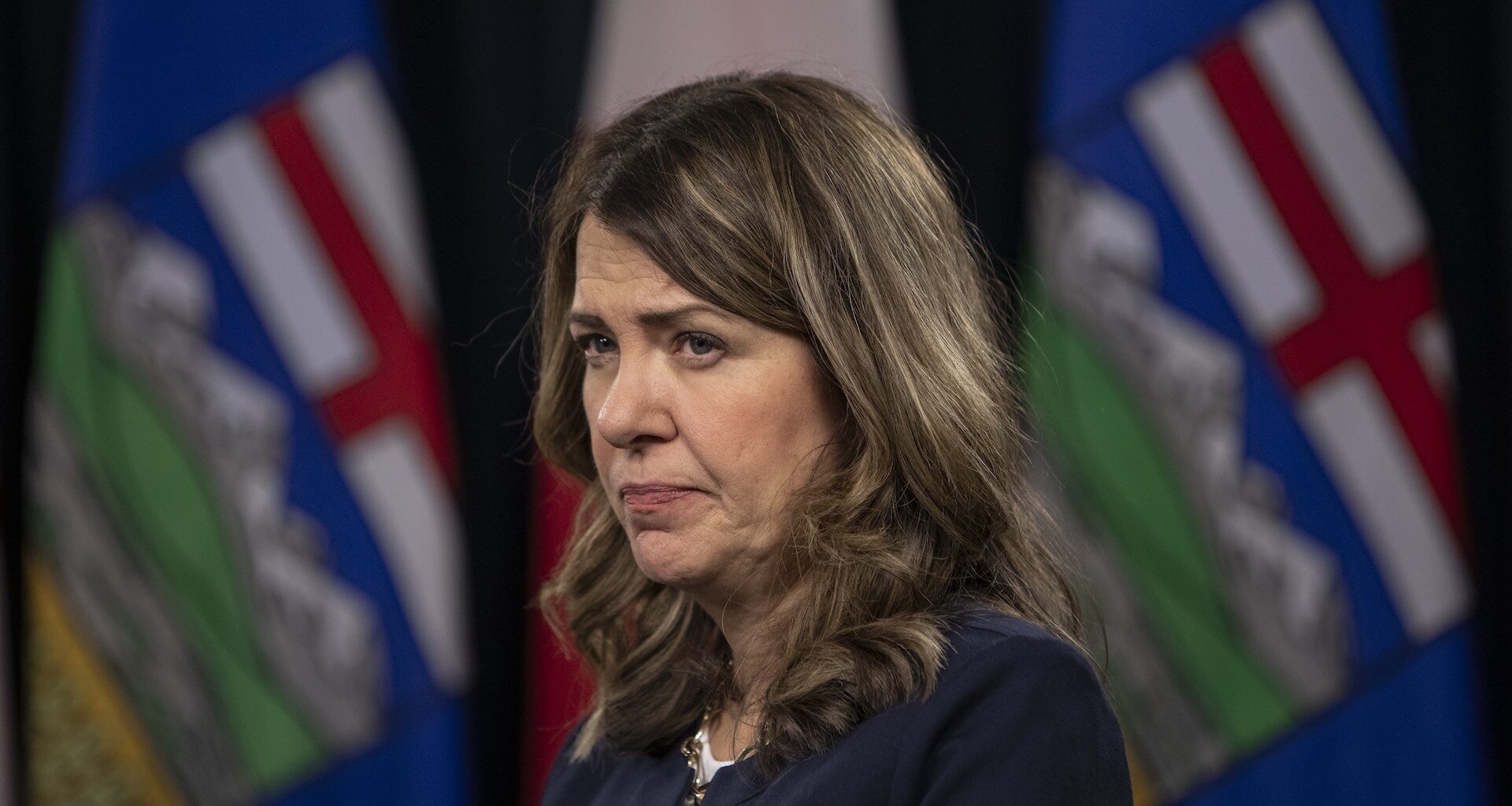 Danielle Smith appears in front of blurred Alberta flags, looking serious.