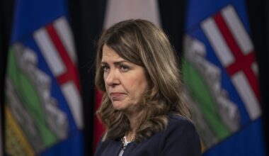 Danielle Smith appears in front of blurred Alberta flags, looking serious.