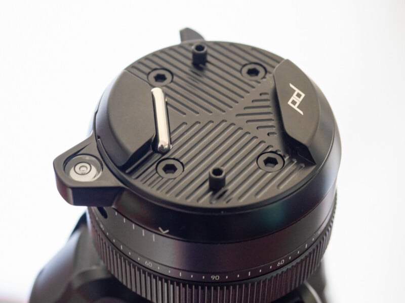 Close-up of a black camera tripod head with textured surface, adjustment knobs, screws, and a metal lever. The "PD" logo is visible on one of the curved locking components.