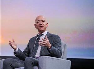 A bald white man in a suit sits in an armchair in front of a pastel colored screen. He is holding both his hands in the air in front of him.