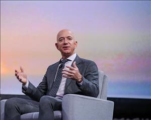 A bald white man in a suit sits in an armchair in front of a pastel colored screen. He is holding both his hands in the air in front of him.